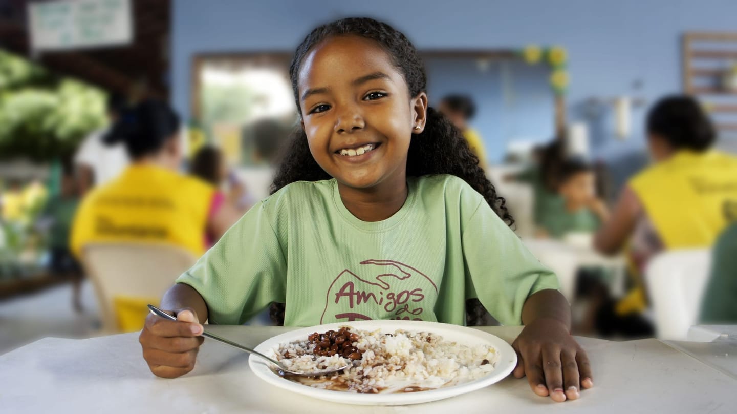 Criança sorrindo com prato de comida no refeitório dos Amigos do Bem